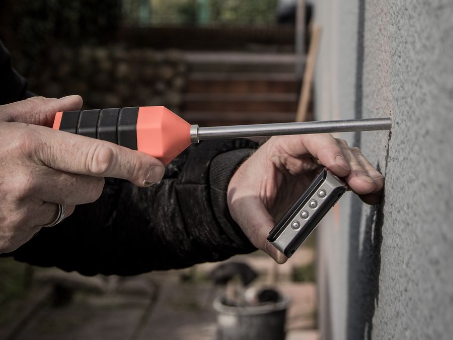 Zwei Hände halten eine Sonde und ein Messgerät an eine verputzte Hauswand